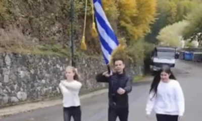 Τρία άτομο περπατούν σε δρόμο κρατώντας ελληνική σημαία