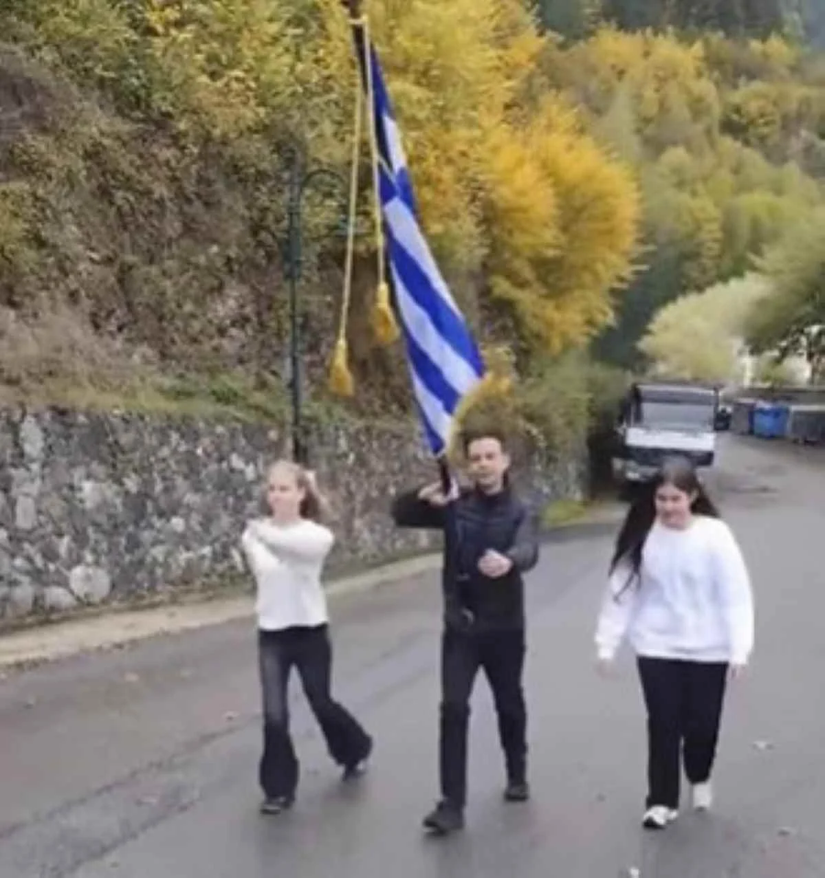 Τρία άτομο περπατούν σε δρόμο κρατώντας ελληνική σημαία