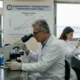 Επιστήμονες με λευκές ποδιές και γάντια εργάζονται σε εργαστήριο πάνω σε μικροσκόπιο και δοκιμαστικούς σωλήνες.