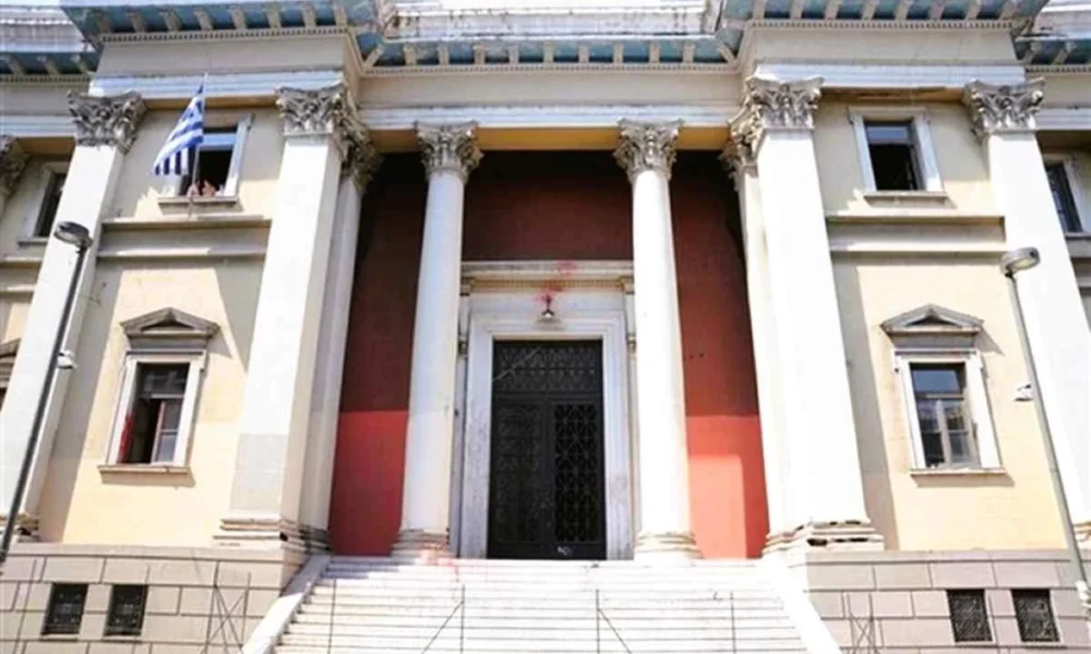 Η πρόσοψη κτηρίου με κίτρινους τοίχους, λευκούς κίονες, μεγάλη μαύρη πόρτα, σκάλες στην είσοδο και μια ελληνική σημαία σε παράθυρο