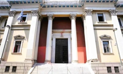 Η πρόσοψη κτηρίου με κίτρινους τοίχους, λευκούς κίονες, μεγάλη μαύρη πόρτα, σκάλες στην είσοδο και μια ελληνική σημαία σε παράθυρο