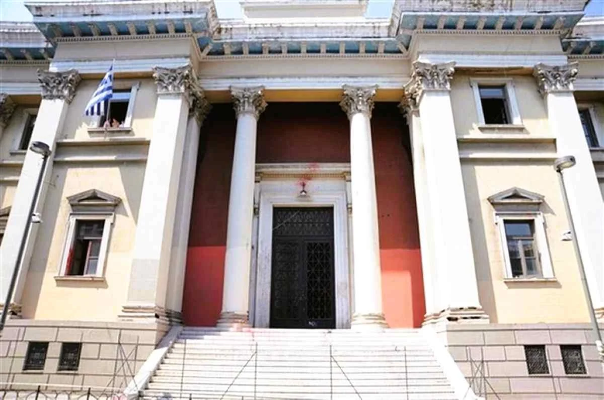 Η πρόσοψη κτηρίου με κίτρινους τοίχους, λευκούς κίονες, μεγάλη μαύρη πόρτα, σκάλες στην είσοδο και μια ελληνική σημαία σε παράθυρο