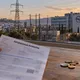 Χέρι κρατά λογαριασμό ρεύματος σε τραπέζι με κέρματα και φλιτζάνι, με θέα κτίριο με λογότυπο και πυλώνες ηλεκτρικής ενέργειας.
