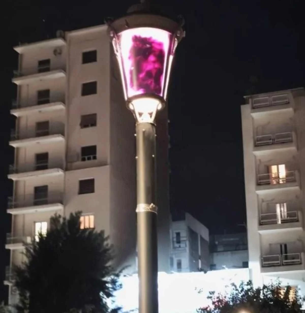 Φωτιστικός στύλος με μουτζουρωμένο φως τη νύχτα και πολυκατοικίες στο βάθος.
