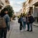 Νεαρή κοπέλα με σακίδδιο πλάτης περπατά μόνη της σε δρόμο, ενώ μια ομάδα τεσσάρων ατόμων στέκεται παραπέρα και ψιθυρίζει.