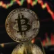 Χρυσά κέρματα Bitcoin μπροστά από οθόνη με κόκκινα και πράσινα διαγράμματα αγοράς