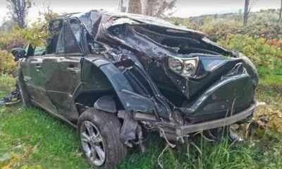 Τρακαρισμένο μαύρο αυτοκίνητο με σοβαρές ζημιές στην οροφή και το πίσω μέρος, ακινητοποιημένο σε αγροτική περιοχή με πράσινο και δ