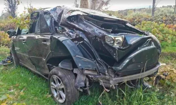 Τρακαρισμένο μαύρο αυτοκίνητο με σοβαρές ζημιές στην οροφή και το πίσω μέρος, ακινητοποιημένο σε αγροτική περιοχή με πράσινο και δ