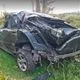 Τρακαρισμένο μαύρο αυτοκίνητο με σοβαρές ζημιές στην οροφή και το πίσω μέρος, ακινητοποιημένο σε αγροτική περιοχή με πράσινο και δ