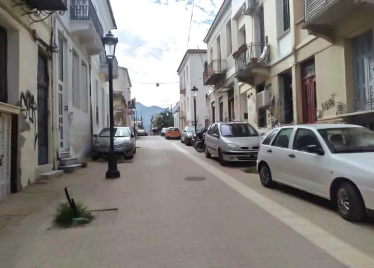 Άποψη στενού δρόμου ανάμεσα σε κτήρια με παρκαρισμένα αυτοκίνητα δεξιά και αριστερά.