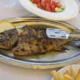 Ψητό ψάρι σε μεταλλική πιατέλα με λεμόνι, συνοδευόμενο από πατάτες τηγανητές και σαλάτα σε τραπέζι