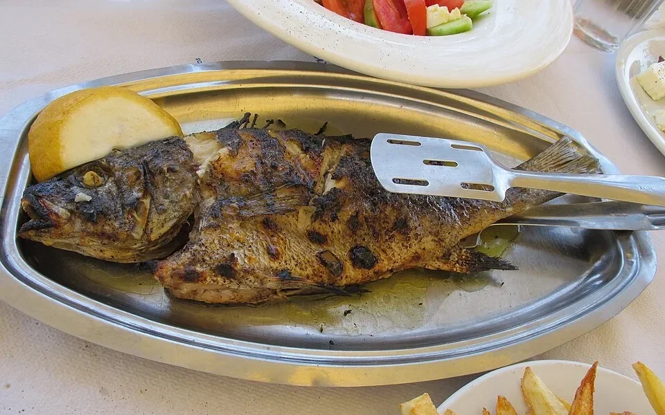Ψητό ψάρι σε μεταλλική πιατέλα με λεμόνι, συνοδευόμενο από πατάτες τηγανητές και σαλάτα σε τραπέζι