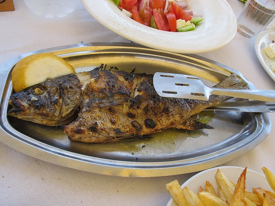 Ψητό ψάρι σε μεταλλική πιατέλα με λεμόνι, συνοδευόμενο από πατάτες τηγανητές και σαλάτα σε τραπέζι