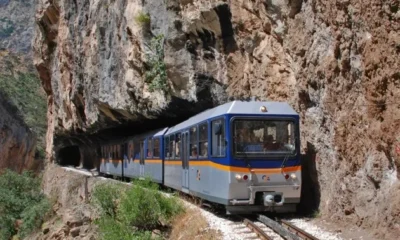 Τρένο κινείται στις ράγες ανάμεσα σε βραχώδη πλαγιά και φυσικό τοπίο.