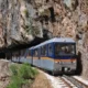 Τρένο κινείται στις ράγες ανάμεσα σε βραχώδη πλαγιά και φυσικό τοπίο.