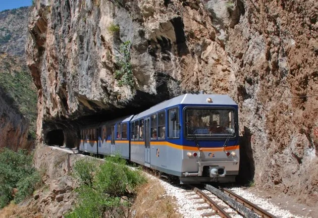 Τρένο κινείται στις ράγες ανάμεσα σε βραχώδη πλαγιά και φυσικό τοπίο.